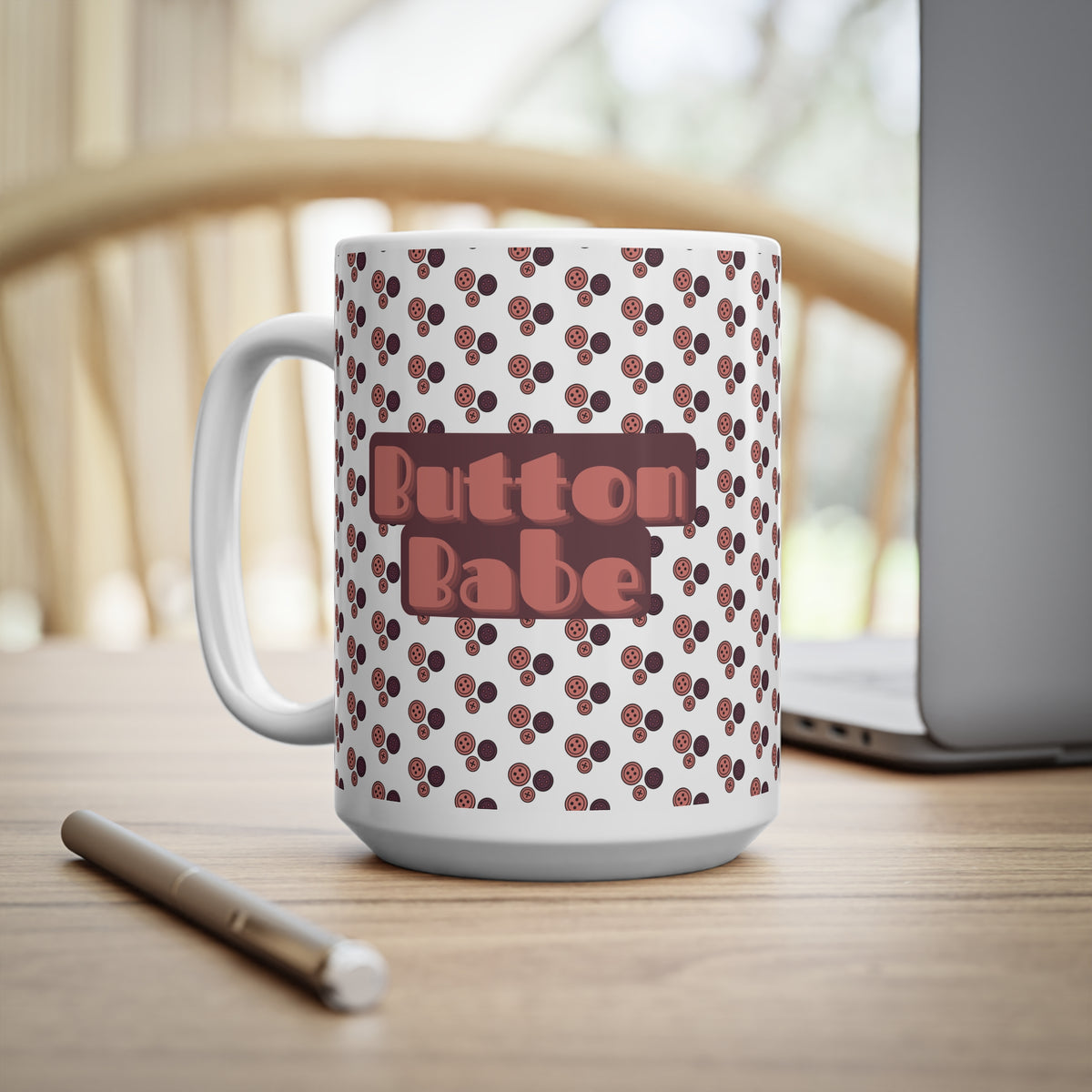 A white ceramic mug with a brown cherry pattern and a pinkish-brown 'Button Babe' text graphic, sitting on a wooden desk next to a laptop and pen.