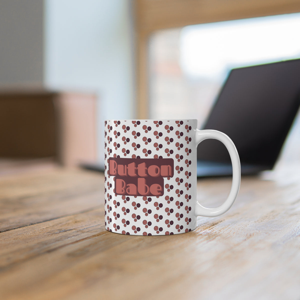 A white ceramic coffee mug with a brown cherry pattern and a central brown label reading 'Button Babe' in pink letters, sitting on a wooden table with a blurred laptop in the background.